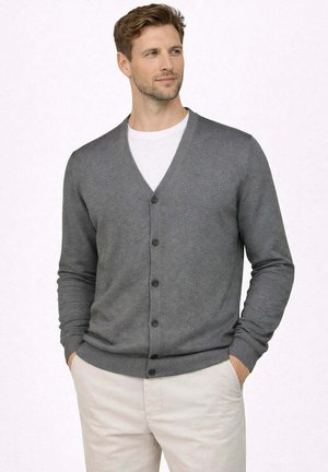 Uomo con capelli castano chiaro che indossa un cardigan grigio abbottonato sopra una camicia bianca e pantaloni beige, con le mani nelle tasche, che guarda leggermente a destra.