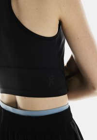 Crop top de sport noir avec une texture lisse et un design minimaliste. Présente un logo sur le côté et une ceinture bleue contrastante en dessous.