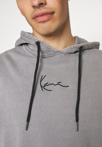 Grauer Kapuzenpulli mit Kordelzug, versehen mit schwarzen Akzenten und gesticktem Logo auf der Brust. Weicher Stoff, lässiger Schnitt mit Kapuze.