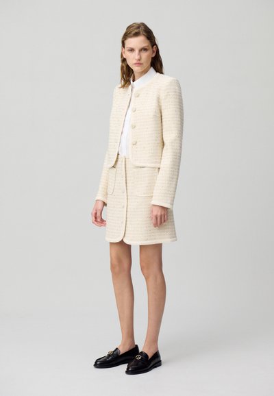 Costume en tweed crème avec une veste ajustée dotée de boutons ronds et d'une jupe assortie. Accentué d'une chemise blanche lisse et de mocassins noirs.