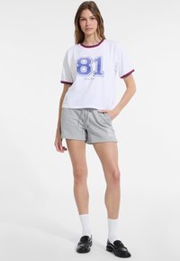 Valkoinen lyhythihainen T-paita, jossa on sininen "81" -printti ja punaiset reunat, yhdistettynä harmaisiin nyöritettävissä shortseihin, joissa on logo. Mustat loafers-kengät ja valkoiset sukat.