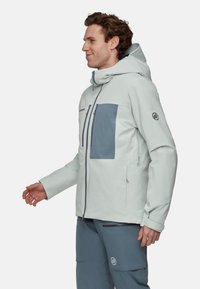 Veste imperméable gris clair avec une poche poitrine gris foncé, capuche et détails à fermeture éclair. Présente une texture lisse et un design ajusté.