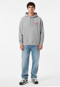 Felpa grigia con tasca frontale e testo rosso. Paio di jeans azzurri chiari, sneakers bianche e nere. Design semplice, texture morbida.