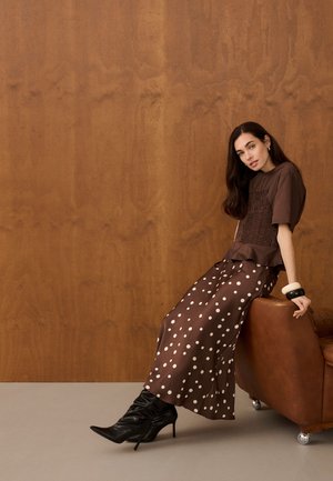 Femme aux longs cheveux foncés, portant un haut marron et une jupe à pois, assise sur une chaise en cuir marron, avec des bottes noires à talons et de grands bracelets.