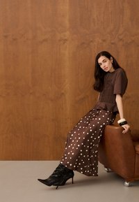 Femme aux longs cheveux foncés, portant un haut marron et une jupe à pois, assise sur une chaise en cuir marron, avec des bottes noires à talons et de grands bracelets.