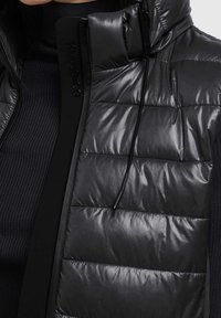 Czarna puchowa kamizelka z błyszczącym pikowanym materiałem, wysokim kołnierzem i czarnymi akcentami. Charakteryzuje się eleganckim designem i regulowanym ściągaczem przy szyi.