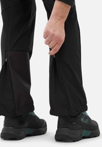 Pantalons noirs d'extérieur avec des côtés zippés, design ajusté et tissu texturé. Associés à des chaussures robustes noires et teal.