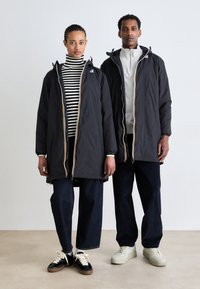 Sorte lange jakker med gule og hvide accenter, båret over en stribet turtleneck og en grå zip-up, parret med mørke jeans og sneakers.