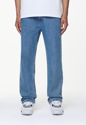Mann trägt locker sitzende hellblaue Jeans aus Denim und weiße Sneakers, steht vor einem schlichten weißen Hintergrund.