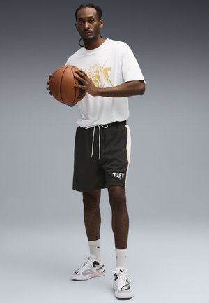 Giocatore di basket con una maglietta bianca con grafica dorata, pantaloni neri con strisce laterali e sneakers bianche. Tiene una pallacanestro.