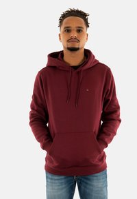 Bordeaux hoodie van zacht materiaal, met een kangoeroezak, capuchon met trekkoord en een klein logo op de borst. Gecoupleerd met lichtdenim jeans.