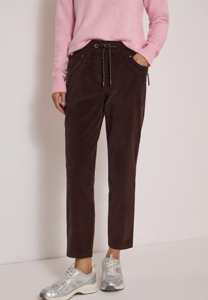 Personne portant un pantalon marron foncé à cordon, un haut rose à manches longues et des baskets argentées, debout contre un mur clair uni.