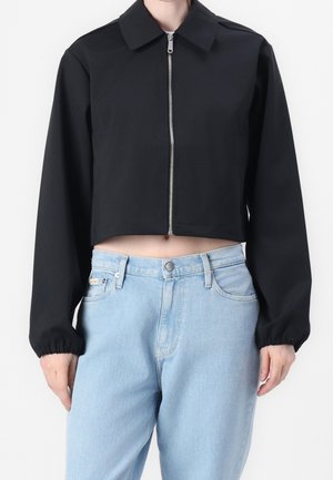 Osoba w czarnej, krótkiej kurtce z długim rękawem i przednim zamkiem błyskawicznym oraz jasnoniebieskich, wysokich dżinsach na białym, jednolitym tle.