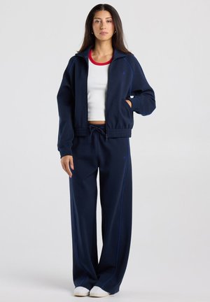 Vrouw staat, draagt een marineblauw trainingspak met een wit cropped T-shirt met een rode kraag en witte slip-on schoenen.