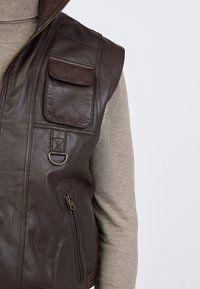 Brauner Lederweste mit hohem Kragen, Reißverschluss, Taschen auf der Vorderseite und dekorativen Nähten. Solide Textur, taillierter Schnitt.