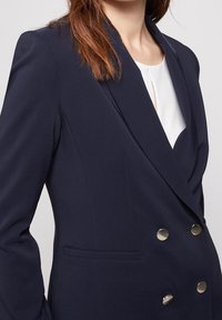 Blazer doppiopetto blu navy con un design elegante, caratterizzato da bottoni di tonalità argento, colletto a revers, tasche frontali e tessuto testurizzato.