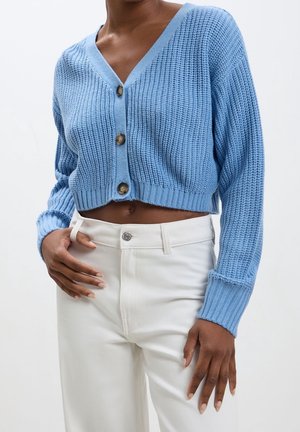 Femme portant un cardigan court en maille bleu clair avec des boutons et un pantalon blanc taille haute, debout devant un fond uni.