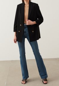 Blazer negro de doble botonadura con botones dorados, combinado con jeans flare azules de tiro medio con un acabado texturizado, y sandalias negras con tiras.