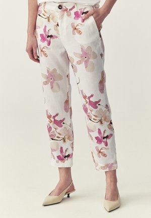 Donna che indossa pantaloni bianchi corti con stampa floreale rosa e beige e scarpe slingback beige con tacco basso, in piedi con una mano in tasca.