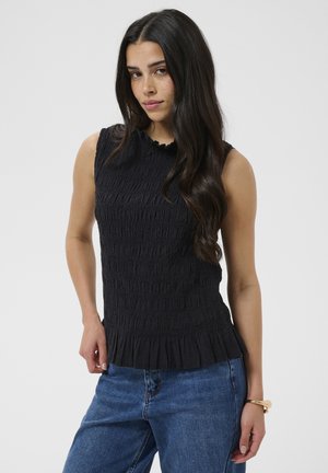 Žena s dlhými tmavými vlasmi, oblečená v čiernom širšom textúrovanom tielku a modrých džínsoch, pózujúca s rukou na boku na jednolatom pozadí.