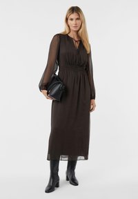 Braunes Langarmkleid mit geraffter Taille, transparenten Ärmeln und einem Bindegürtel am Hals, kombiniert mit schwarzen Absatzstiefeln und einer kleinen schwarzen Tasche.