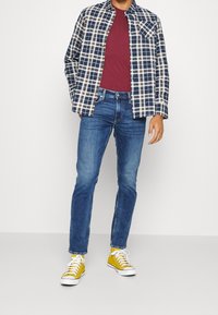 Rutig skjorta i marinblått, vitt och brunt över en vinröd t-shirt, kombinerat med blå jeans och gula sneakers.