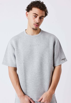 Giovane uomo con capelli ricci che indossa una felpa oversize a maniche corte grigio chiaro su uno sfondo uniforme.