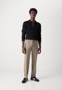 Polo en tricot noir avec fermeture éclair, pantalon beige à cordon de serrage et baskets noires avec rayures blanches. Texture lisse avec coupe décontractée.