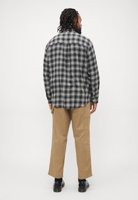 Man met dreadlocks, gekleed in een zwart-wit geruit overhemd, beige broek met zijzakken, zwarte sokken en zwarte schoenen, staand met zijn rug naar de camera toe.