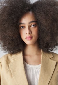 Blazer en lin beige avec des revers, associé à un haut léger à côtes. Les cheveux sont volumineux et bouclés, encadrant le visage.