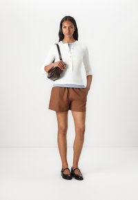 Hvit ribbet langermet topp lagd over en blåstripet skjorte, sammen med brune shorts og svarte lakkflats; holder en brun veske.
