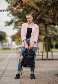 Čern�ý batoh s barevnými tečkami, rolovací design a nastavitelné popruhy. Modelka má na sobě růžový blazer a roztrhané džíny.