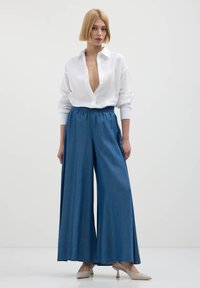 Camicia bianca con colletto a V profondo, maniche arrotolate; pantaloni in denim a gamba larga blu con cintura elastica; tacchi leggeri beige completano l'outfit.