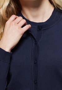 Tmavě námořnický modrý svetr na zapínání s kulatým výstřihem, texturovanou látkou a čtyřmi viditelnými knoflíky. Detailní záběr na ruku u výstřihu.