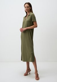 Abito midi verde oliva con colletto, maniche corte e spacchi laterali. Realizzato in tessuto morbido con una vestibilità comoda. Indossato con sandali alti color sabbia.