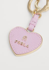 Privjesak u obliku srca napravljen od ružičaste teksturirane kože s metalnom bordurom u zlatnoj boji. Na prednjoj strani se nalazi urezano "FURLA" brendiranje.