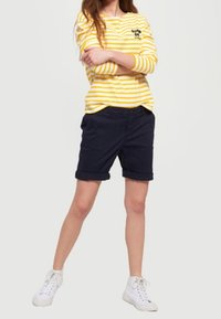 Yellow and white striped long-sleeve shirt with a chest logo, paired with navy rolled-up shorts and white sneakers.
