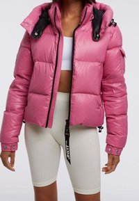 Rosa puffjacka med glansig yta, med en tvåfärgad huva i svart, dragkedja och quiltad design. Buren över en vit topp och ljusa ribbade shorts.
