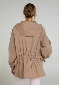 Lekka jasnobeżowa kurtka z kapturem, ściągacz w pasie z możliwością regulacji oraz złotymi detalami, a także luźne rękawy. Gładki, wodoodporny materiał.