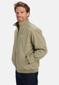Hombre sonriente con cabello castaño claro, vistiendo una chaqueta beige con cierre y con las manos en los bolsillos, de pie frente a un fondo blanco liso.