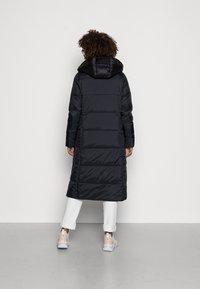Sort puffjakke med hætte, knælang, quiltet design, med elastiske manchetter og en glat, skinnende overflade. Båret over hvide bukser og sko.