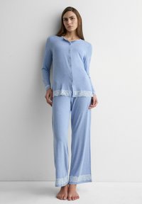 Frau trägt ein hellblaues, langärmliges, geknöpftes Pyjama-Oberteil und passende weite Hosen mit weißer Spitzenborte, steht barfuß an einer weißen Wand.