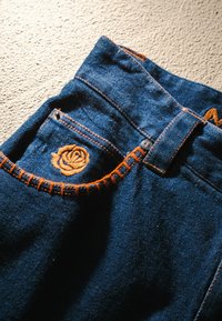 Jeans in denim con tessuto blu scuro, cuciture arancioni e un ricamo di una rosa sulla tasca. Presentano una classica chiusura frontale con bottoni e cerniera.