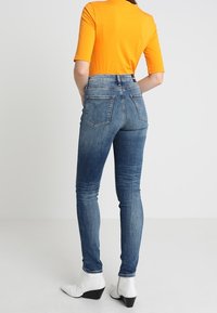 Vaqueros de denim azul de cintura alta con un acabado desgastado, bolsillos traseros y un ajuste ajustado, combinados con una blusa naranja de manga corta y botas blancas hasta el tobillo.