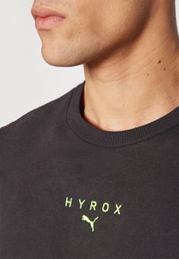 Grå hettegenser med rund hals og ribbestrikkede ermer. Har et grønt "HYROX"-logo og et Puma-logo under teksten. Glatt tekstur.