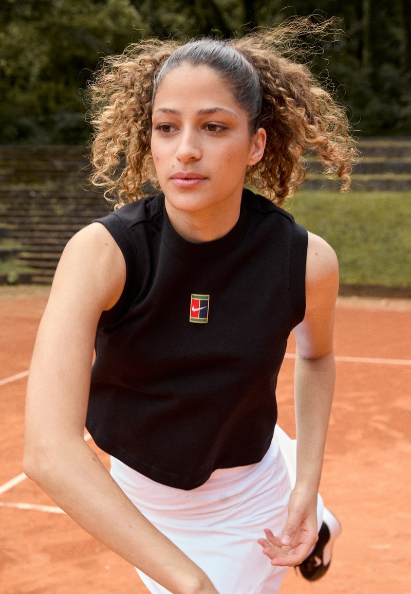 Top corto sin mangas negro con un pequeño y colorido logo de Nike y una falda blanca fluida, colocados en una cancha de tenis de arcilla con vegetación de fondo.