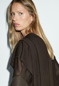Brown, layered knit top with sheer accents, featuring loose sleeves. Model has long, wavy hair and a neutral backdrop.