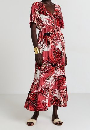 Femme portant une robe maxi à imprimé tropical rouge et blanc avec manches courtes, un bracelet manchette en or et des sandales beiges texturées.