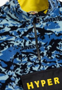 Sudadera de forro polar en azul con patrón abstracto en negro, cuenta con un cuello alto, cremallera frontal y un parche negro con la inscripción "HYPER" en amarillo.