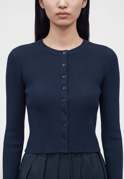 Femme portant un cardigan côtelé bleu marine à manches longues boutonné, associé à une jupe plissée foncée, sur un fond clair uni.